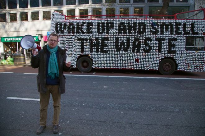 A man with a foghorn and a cup standing in front of a double decker bus with coffee cups on its side that spell out 'Wake up and smell the waste.'