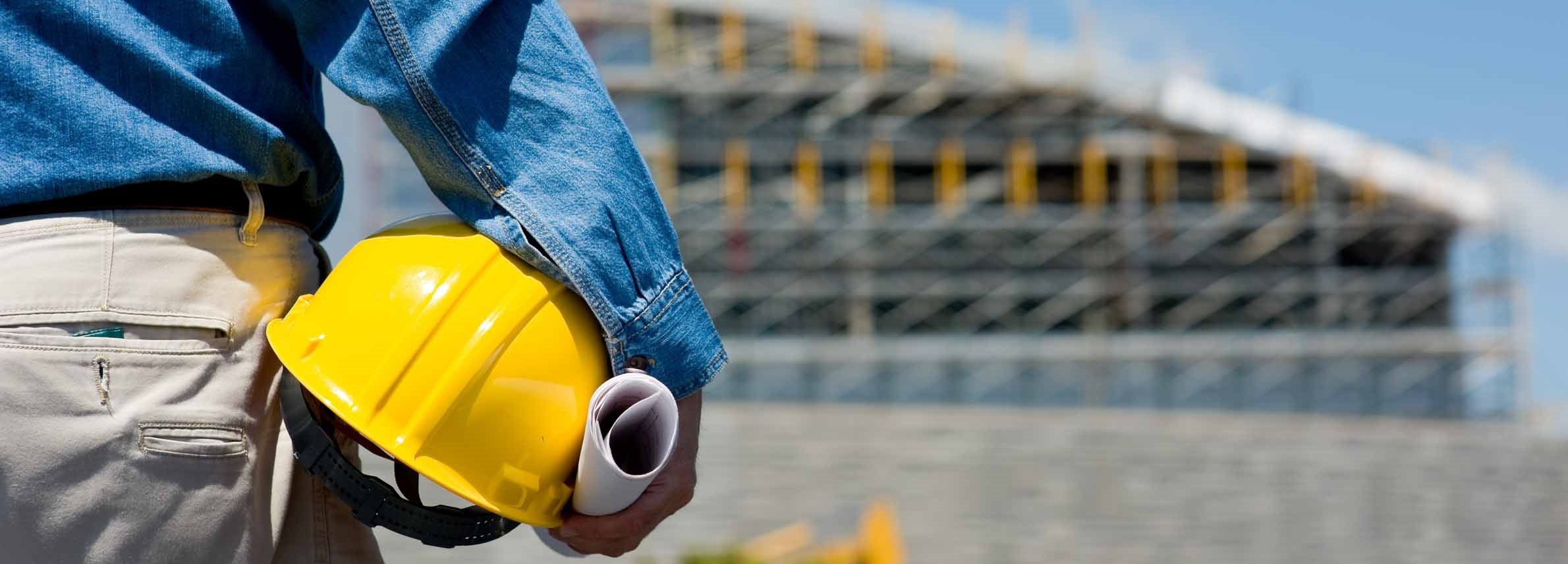 A man looking at a building site holding a hard hat and a rolled-up piece of paper.