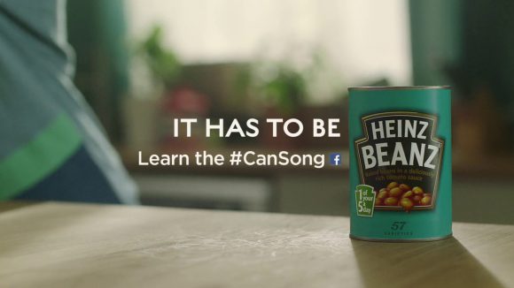 Text which reads "IT HAS TO BE. Learn the #CanSong", alongside a Facebook symbol and a can of Heinz Beanz on a table.