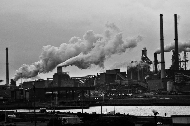 A factory with smoke billowing out it's towers, in black and white.