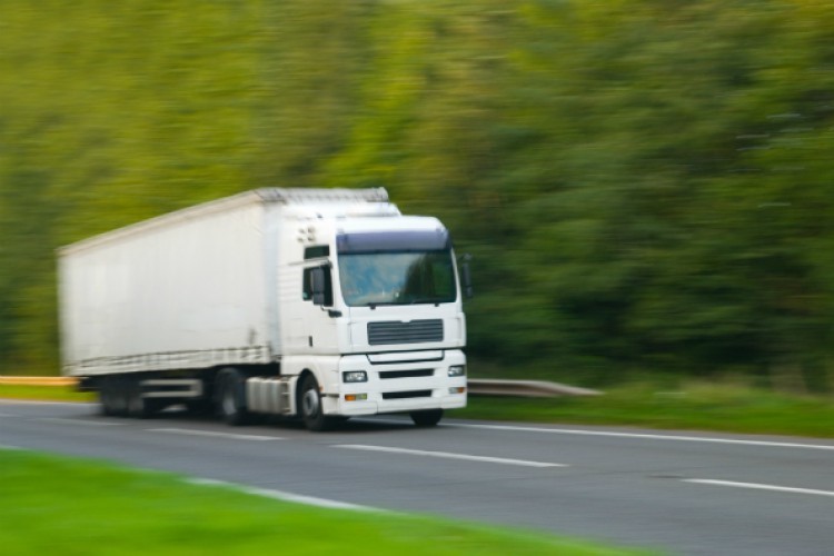 A white HGV driving on a road past trees, slightly blurred to indicate its speed.