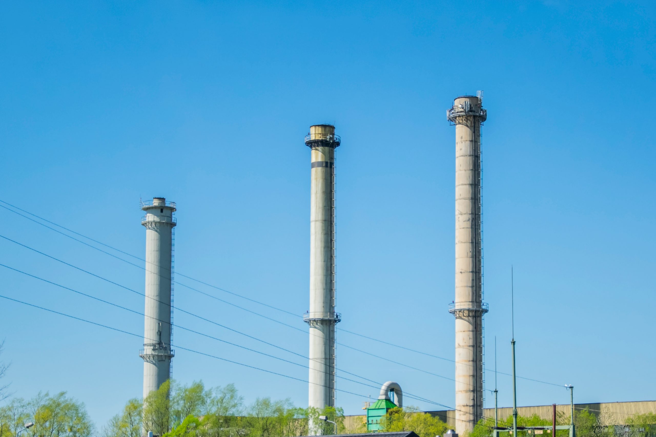 Three tall chimneys that have electric wires passing between the, against a background of a blue sky and greenery.