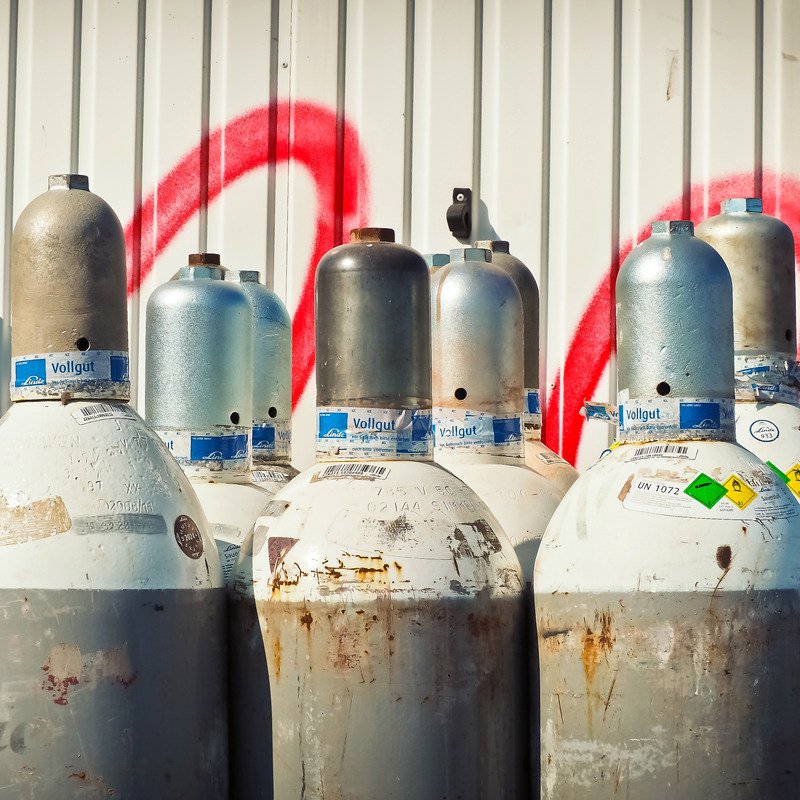 A collection of rusted gas cylinders against a white ridged wall with red spray paint on.