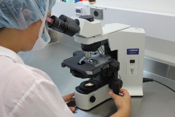 A scientist wear face protection looking into a microscope.