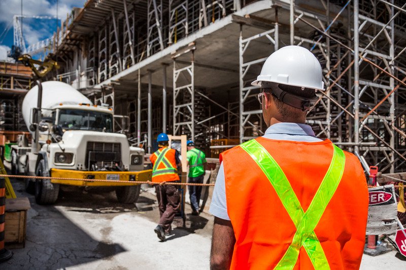 A construction worker in a yellow and orange hi-vis jacket with a hard hat on looking on to a construction site.