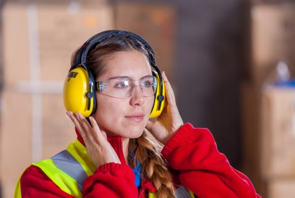 A women wearing a red jacket with a hi-vis vest on top, safety goggles and hearing protection.