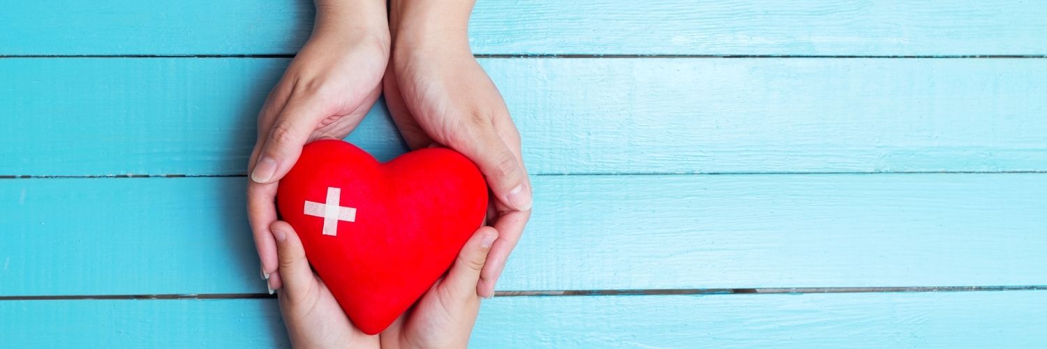 Two sets of hands holding a heart (cartoon shape) with a white cross on it